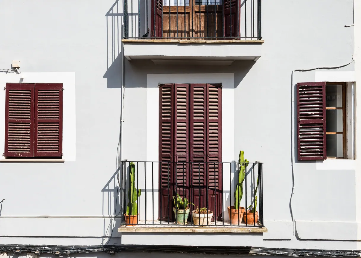 Balcon ensoleillé sur façade blanche avec volets marrons et cactus.