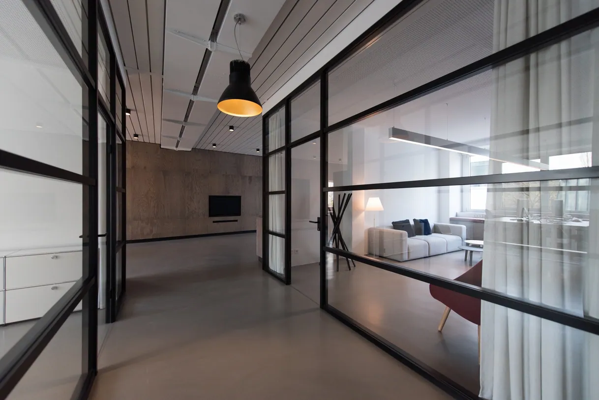Couloir de bureau moderne avec cloisons vitrées noires, salon visible.