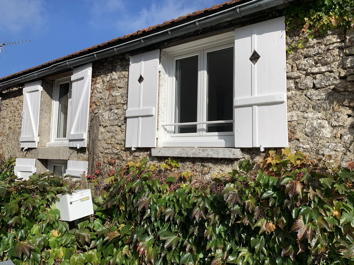 Façade de maison en pierre avec volets blancs et feuillage vert luxuriant.