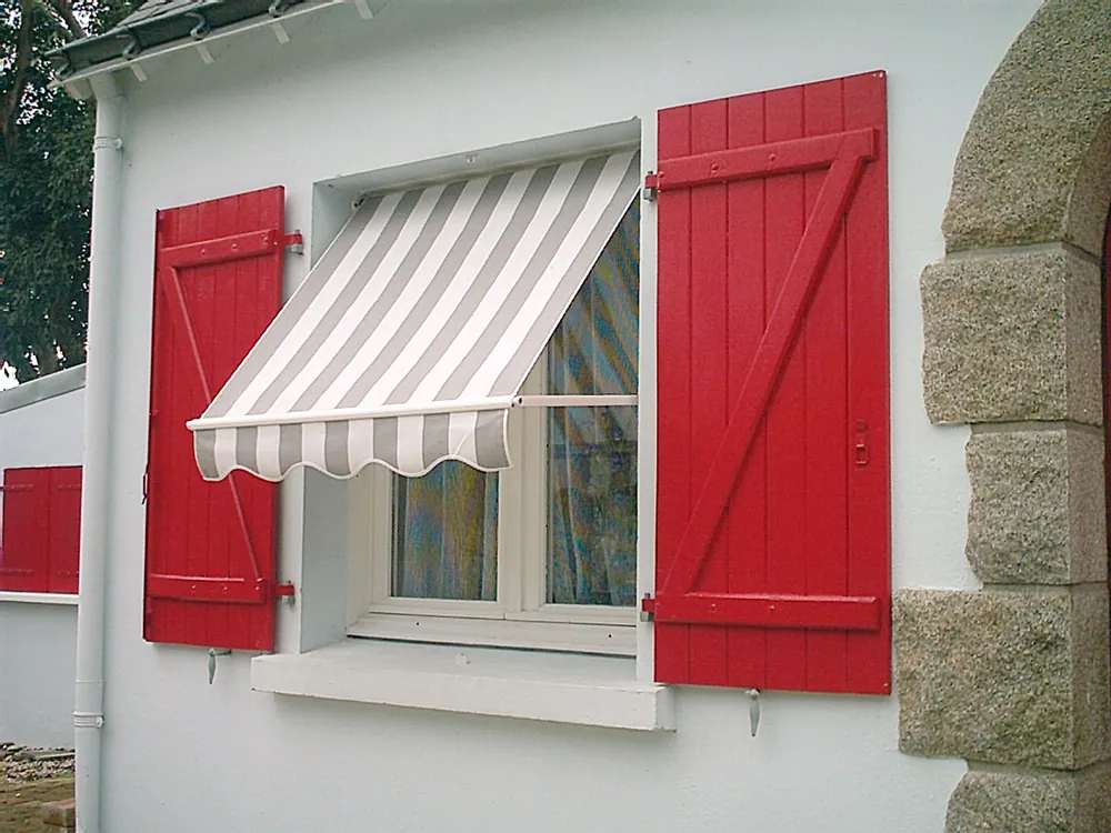 Fenêtre de maison blanche avec volets rouges et auvent rayé blanc et gris.