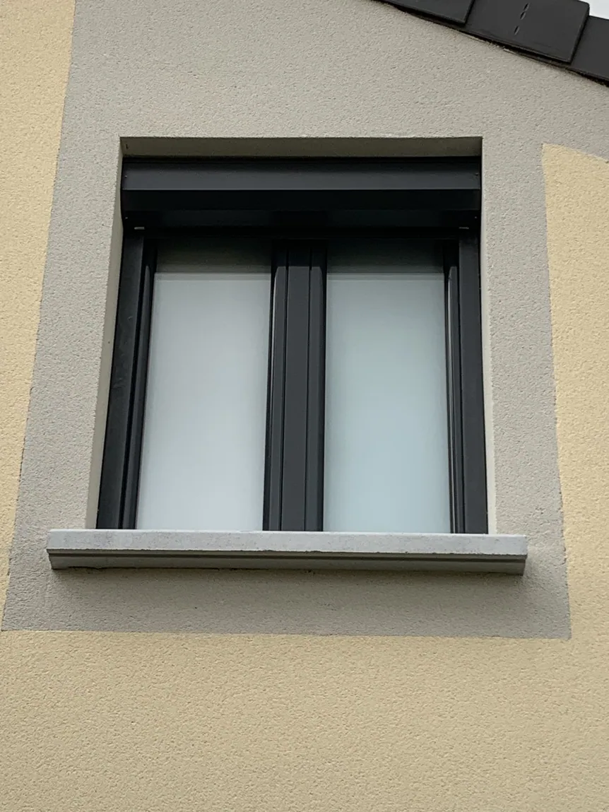 Fenêtre moderne avec volet roulant sombre sur façade en crépi clair.
