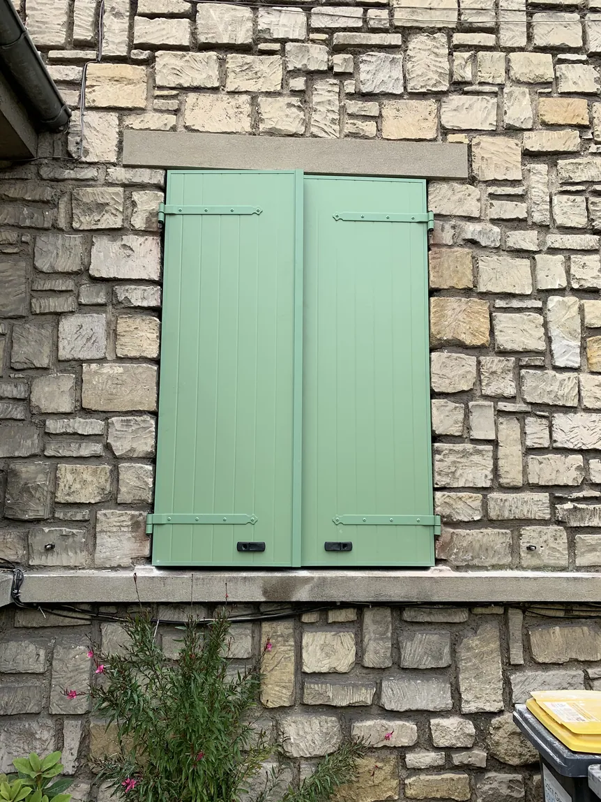Fenêtre avec volets verts clairs fermés sur un mur en pierre rustique.