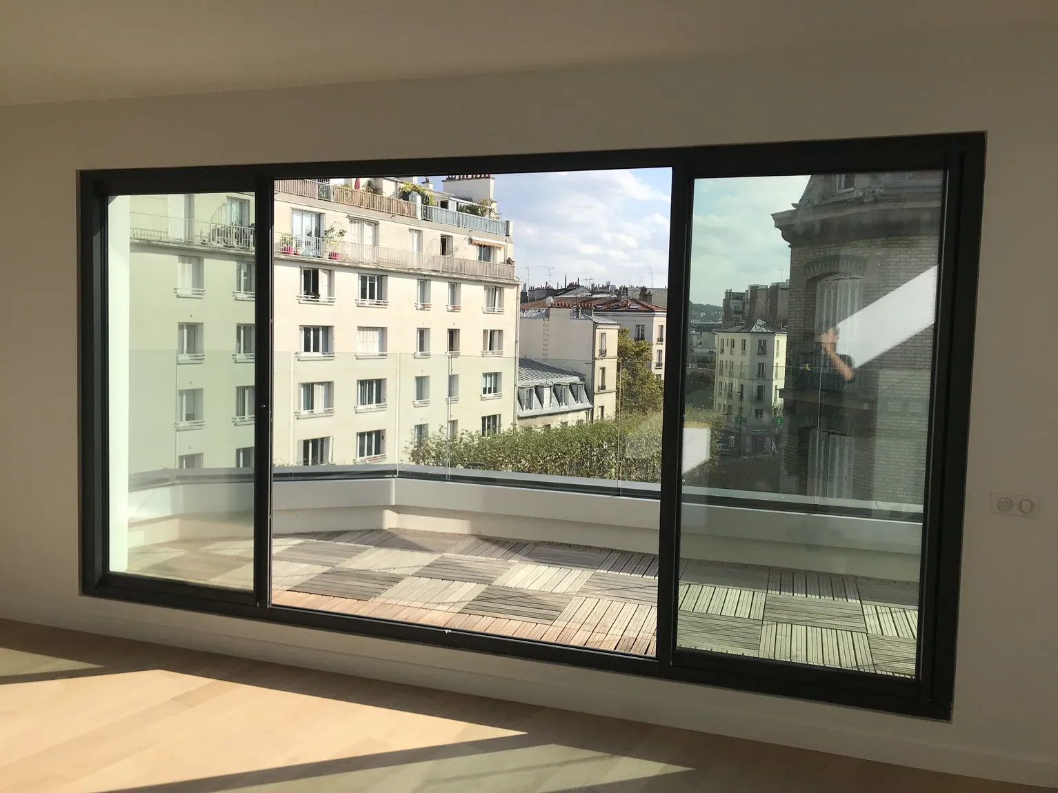 Grande fenêtre moderne avec vue sur terrasse et immeuble urbain.
