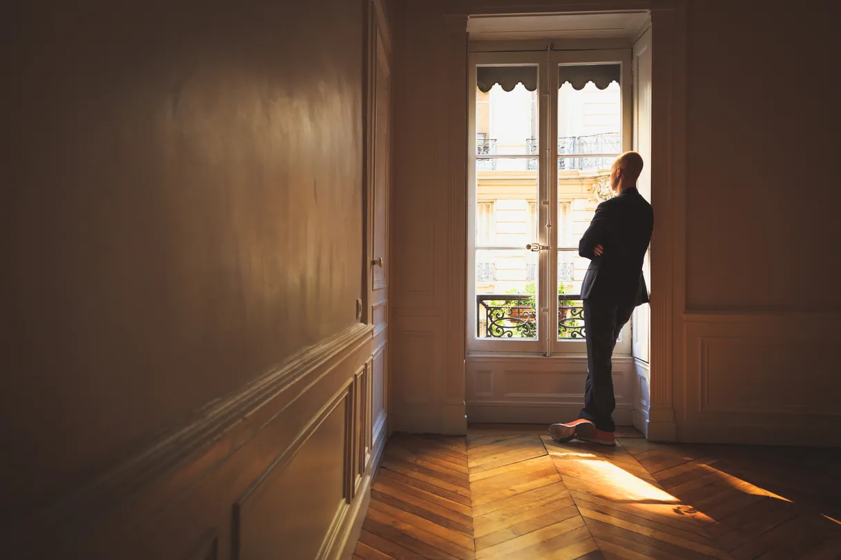 Homme debout dans pièce vide, contemplant la vue par la fenêtre.