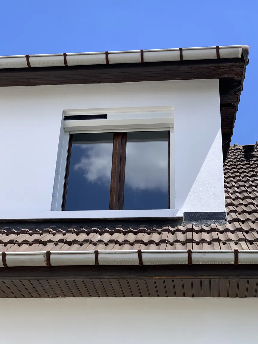 Lucarne blanche avec volet fermé sous ciel bleu sur toiture marron.