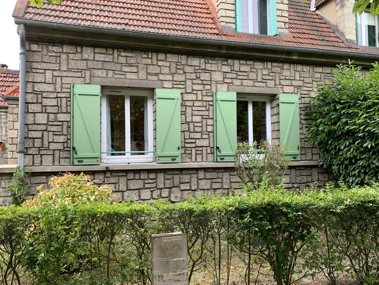 Maison en pierre, fenêtres avec volets verts, toit et buissons.