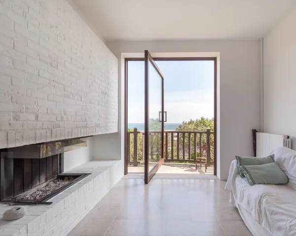 Salon lumineux, cheminée blanche, canapé et porte ouverte sur une vue mer.