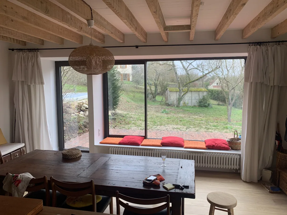 Salle à manger rustique, table en bois, poutres, grande baie vitrée sur jardin.